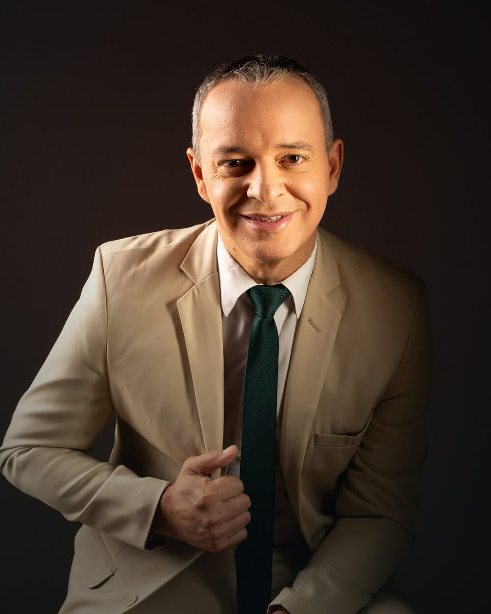 Paulo Crispim sentado, de terno claro e gravata verde, sorrindo para a câmera